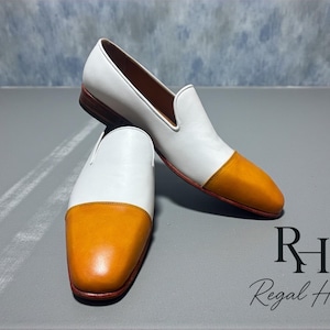 K&ouml;nnte beinhalten: Ein Paar wei&szlig;e und orangefarbene Leder-Loafer. Die Schuhe haben ein wei&szlig;es Obermaterial mit einer orangefarbenen Zehenkappe. Die Sohlen sind braun. Die Schuhe werden auf einer grauen Oberfl&auml;che pr&auml;sentiert.