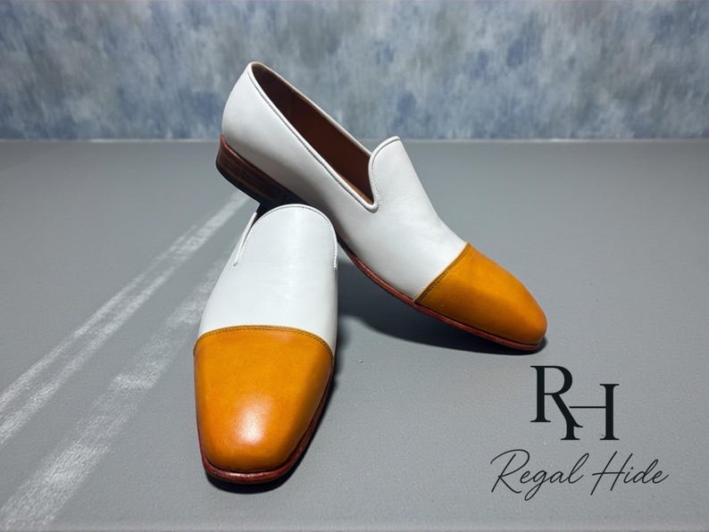 K&ouml;nnte beinhalten: Ein Paar wei&szlig;e und orangefarbene Leder-Loafer. Die Schuhe haben ein wei&szlig;es Obermaterial mit einer orangefarbenen Zehenkappe. Die Sohlen sind braun. Die Schuhe werden auf einer grauen Oberfl&auml;che pr&auml;sentiert.