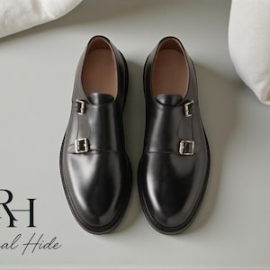 Pode incluir: Um par de sapatos pretos de couro com fivelas duplas e fivelas prateadas. Os sapatos têm um acabamento polido e um design clássico. A imagem também inclui o texto "Regal Hide".
