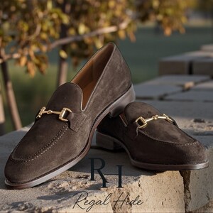 May include: A pair of dark brown suede loafers with gold-colored metal accents. The shoes are displayed on a stone surface with the brand name "Regal Hide" visible. The loafers have a classic design.