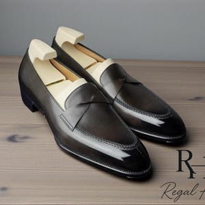 May include: A pair of dark gray leather loafers with a glossy finish. The shoes have a classic design with a low heel and a decorative strap across the top. Shoe trees are inserted inside. The Regal Hide logo is in the bottom right corner.