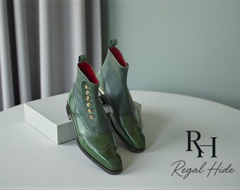 May include: Green leather ankle boots with a classic design. The boots have a button closure, brogue detailing, and a red interior. The boots are displayed on a white surface.