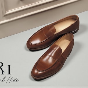 May include: A pair of brown leather loafers with a classic design. The shoes are displayed on a light gray surface, with the "Regal Hide" logo in the lower left corner. The loafers have a polished finish.