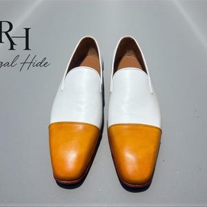 K&ouml;nnte beinhalten: Ein Paar wei&szlig;e und orangefarbene Leder-Loafer. Der obere Teil des Schuhs ist wei&szlig;, w&auml;hrend die Zehenkappe orange ist. Die Schuhe werden auf einer grauen Oberfl&auml;che pr&auml;sentiert. Der Text "Regal Hide" befindet sich in der oberen linken Ecke.