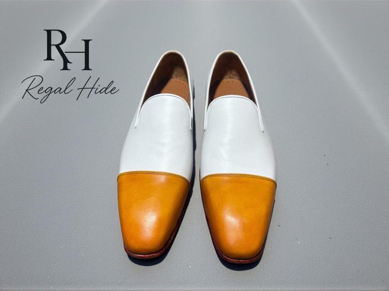 K&ouml;nnte beinhalten: Ein Paar wei&szlig;e und orangefarbene Leder-Loafer. Der obere Teil des Schuhs ist wei&szlig;, w&auml;hrend die Zehenkappe orange ist. Die Schuhe werden auf einer grauen Oberfl&auml;che pr&auml;sentiert. Der Text "Regal Hide" befindet sich in der oberen linken Ecke.