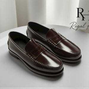 May include: A pair of dark brown leather penny loafers. The shoes have a classic design with a rounded toe, a strap across the vamp, and a low heel. The shoes are displayed on a white surface. The text "RH Regal Hide" is in the background.