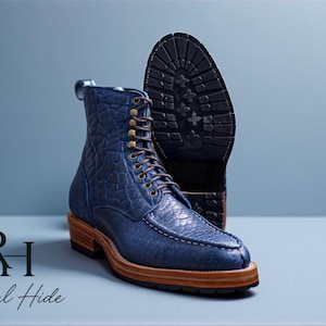 Puede incluir: Botines de cuero azul con un patrón texturizado, cordones marrones y suela de madera. Las botas tienen un diseño robusto y se muestran con la suela de una bota a juego en el fondo. Calzado elegante.