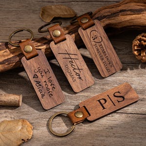 Puede incluir: Llaveros de madera con correas de cuero y anillos de metal. Los llaveros tienen texto grabado, incluyendo nombres, iniciales y frases como "I love you more Alicia" y "3 years down forever to go".