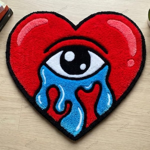 May include: A heart-shaped rug with a red background and a black border. The rug features a white eye with a black pupil and blue tears. Graphic design books and a potted plant are in the background.