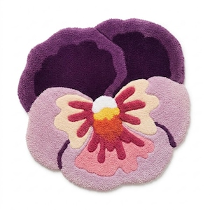 May include: A plush, flower-shaped rug in the form of a pansy. The rug features a deep purple outer petal, a light purple inner petal, and a center with cream, orange, pink, and white accents. The rug is designed to be placed on the floor.