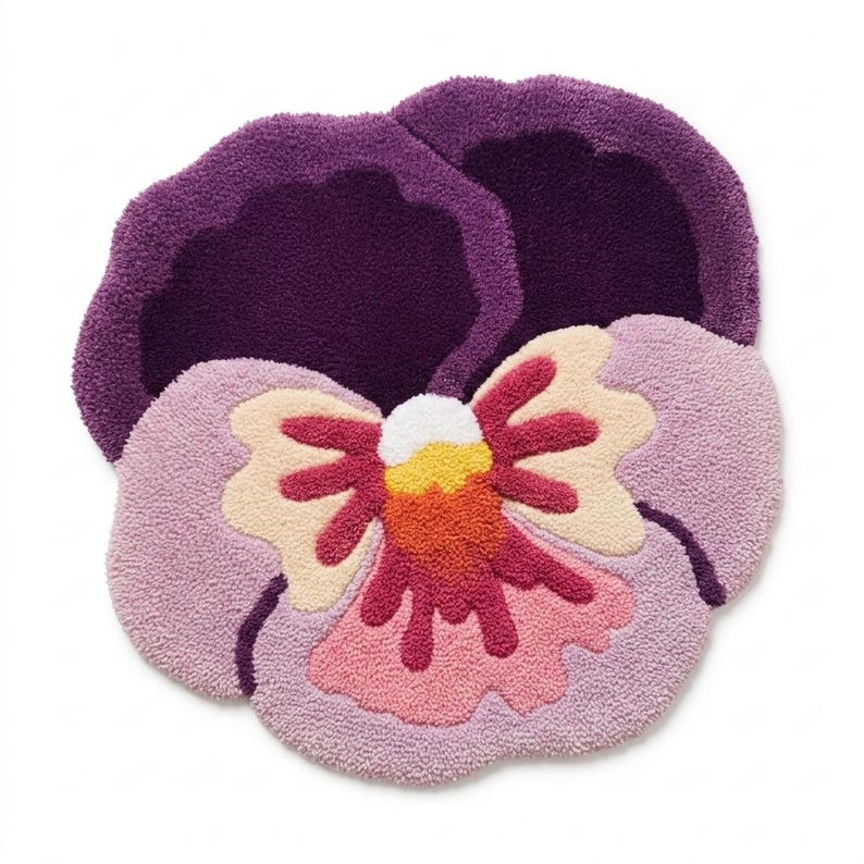 May include: A plush, flower-shaped rug in the form of a pansy. The rug features a deep purple outer petal, a light purple inner petal, and a center with cream, orange, pink, and white accents. The rug is designed to be placed on the floor.