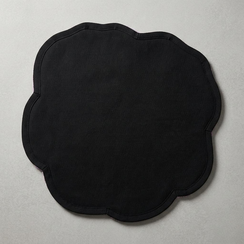 May include: A black placemat with a scalloped edge. The placemat has a solid black surface and a decorative, wavy border. The shape is reminiscent of a cloud or flower, adding a touch of elegance to any table setting.