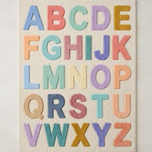 May include: A rectangular off-white rug featuring the English alphabet in raised, tufted letters. Each letter is rendered in a distinct pastel color, including shades of blue, pink, teal, purple, yellow, and orange, arranged in five rows of five letters.