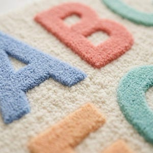 May include: A tufted textile wall hanging or rug featuring the letters A, B, C, and D in pastel colors. The letter A is blue, B is coral, C is mint green, and D is peach, all set against a cream-colored, textured, looped pile background.