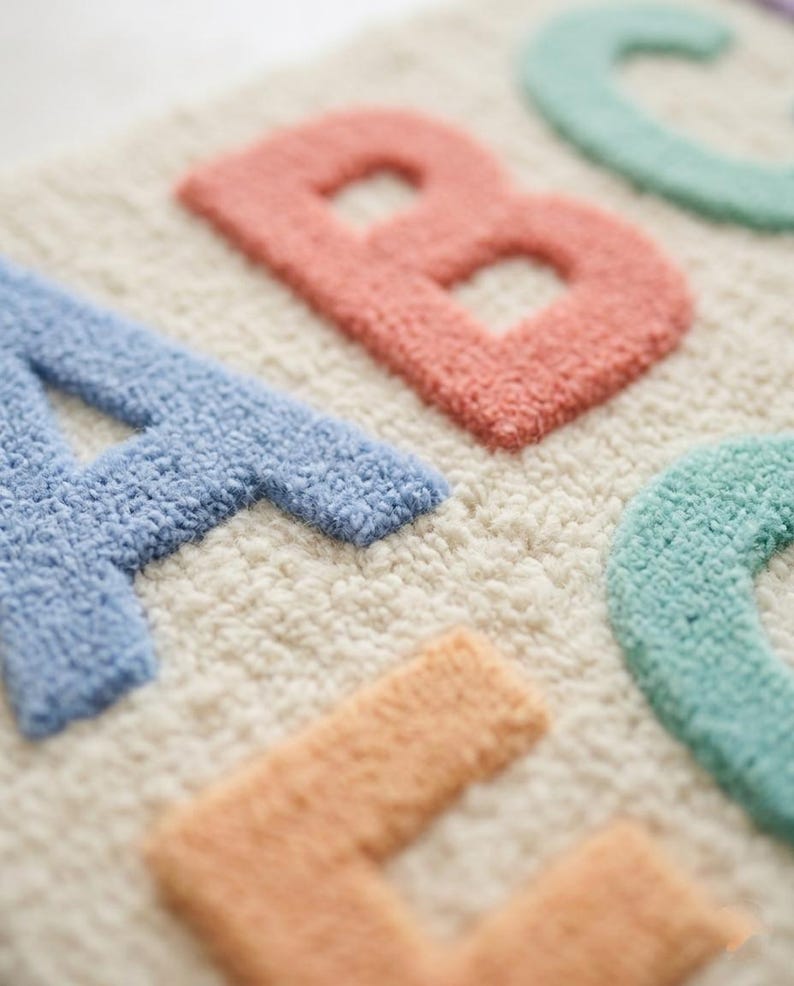 May include: A tufted textile wall hanging or rug featuring the letters A, B, C, and D in pastel colors. The letter A is blue, B is coral, C is mint green, and D is peach, all set against a cream-colored, textured, looped pile background.
