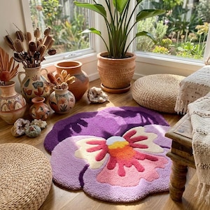 May include: A purple, pink, and yellow flower-shaped rug sits on a wooden floor. The rug has a textured, tufted design. Decorative vases, a potted plant, and woven poufs surround the rug, creating a cozy, bohemian aesthetic.