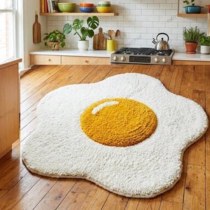 May include: A white, fluffy rug shaped like a fried egg with a yellow yolk center. The rug is on a hardwood floor in a kitchen setting. The kitchen has wooden cabinets, a stove, and various kitchen items.