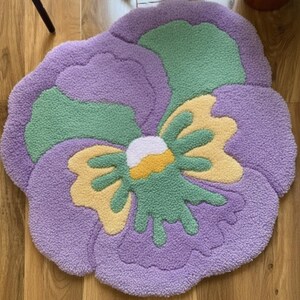 May include: A plush, flower-shaped rug in shades of purple, green, yellow, and white. The rug's design mimics a pansy, with soft, textured petals and a central flower detail. The rug is placed on a wooden floor.