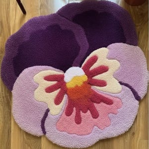 May include: A handmade, plush rug in the shape of a pansy flower. The rug features a deep purple top petal, with light purple, pink, cream, and red petals. The center of the flower is yellow and white.