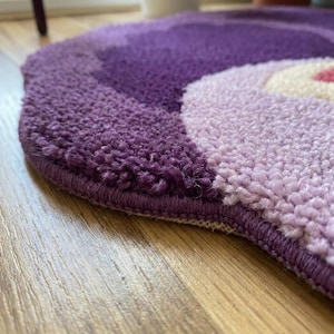 May include: A round, tufted rug in shades of purple, lavender, and cream. The rug has a textured surface and a stitched border. The design features a swirl pattern with a pink accent in the center. The rug is placed on a wooden floor.
