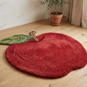 May include: A red apple-shaped rug with a green leaf and brown stem. The rug is made of a soft, textured material and is placed on a wooden floor. The rug measures approximately 100 cm in diameter.