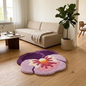 May include: A plush, flower-shaped rug in shades of purple, pink, and yellow. The rug is placed on a hardwood floor in a living room setting. The flower design has a detailed, textured appearance, adding a decorative element to the room.