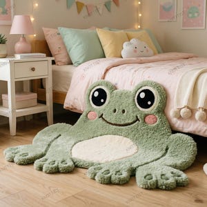 May include: A plush, sage green frog-shaped rug featuring large black and white eyes, a smiling mouth, and pink cheeks. The rug has a cream-colored belly and textured, tufted fabric, positioned on a light wood floor in front of a bed.