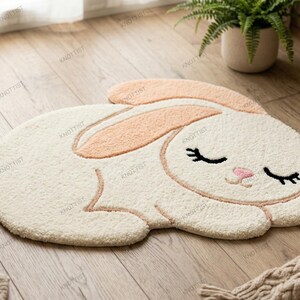 May include: A cream-colored area rug shaped like a sleeping rabbit with long peach-colored ears, a small pink nose, and closed eyes with black eyelashes. The rug features a soft, tufted texture and a small peach-colored tail on the left side.