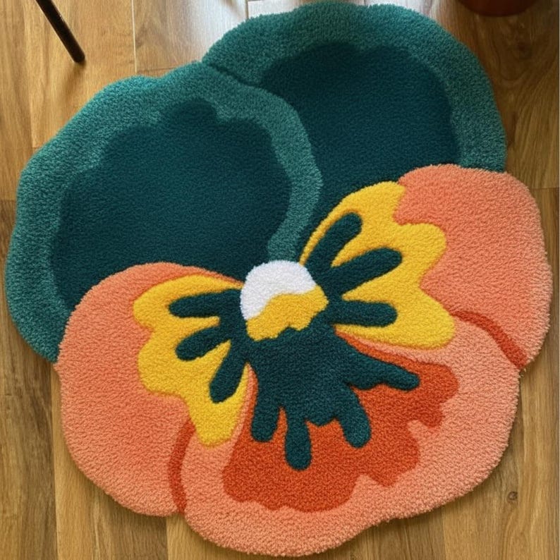 May include: A plush, flower-shaped rug with a vibrant design. The rug features a large, stylized pansy with petals in shades of orange, yellow, and green. The center of the flower is white. The rug is placed on a wooden floor.