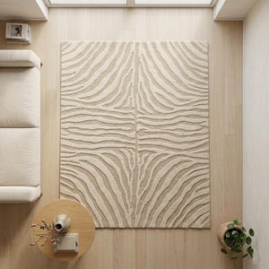 Puede incluir: Una alfombra rectangular con una textura de pelo alto y bajo que presenta un patrón repetitivo de rayas de cebra en color beige y crema. El diseño consiste en líneas orgánicas y fluidas que crean un efecto visual simétrico en toda la superficie.