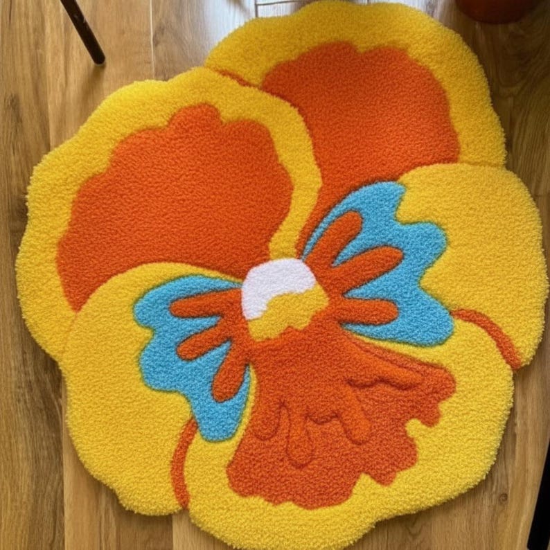 May include: A colorful, flower-shaped rug with a textured, tufted design. The rug features a vibrant color palette of yellow, orange, blue, and white. The rug is designed to resemble a flower, with petals and a central design.