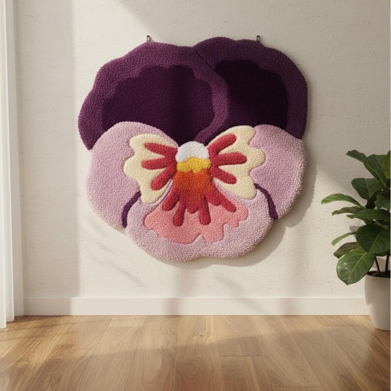 May include: A large, plush, purple, pink, and cream-colored pansy flower wall hanging. The flower is mounted on a white wall above a wooden floor. The flower's petals are a mix of light pink and purple, with a cream and red center.