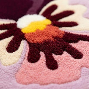 May include: Close-up of a colorful, textured flower design. The flower features petals in shades of pink, cream, and burgundy, with a yellow and orange center. The design is likely part of a rug or textile.