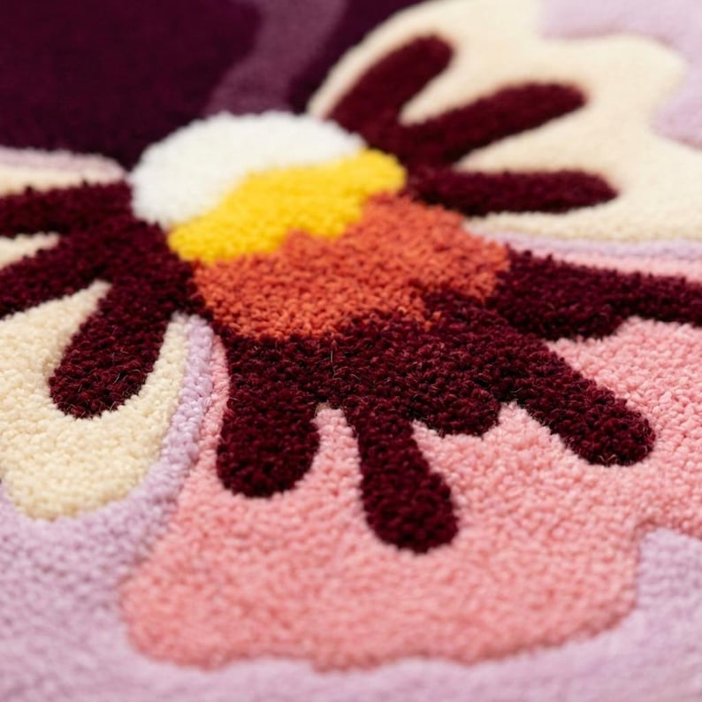 May include: Close-up of a colorful, textured flower design. The flower features petals in shades of pink, cream, and burgundy, with a yellow and orange center. The design is likely part of a rug or textile.