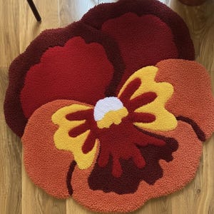 May include: A flower-shaped rug with a vibrant design. The rug features a large, multi-layered flower in shades of red, orange, and yellow. The center of the flower is white. The rug is made of a soft, textured material.