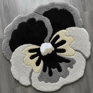 May include: A handmade, flower-shaped rug with a textured, tufted design. The rug features a black center, surrounded by light gray, dark gray, and beige petals. The rug is a decorative floor covering.