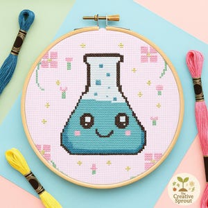 May include: A cross-stitch embroidery hoop with a smiling, cartoon-style Erlenmeyer flask in blue and black on a pink background. The design includes pink flowers and yellow accents. Embroidery threads in blue, pink, and yellow are visible.