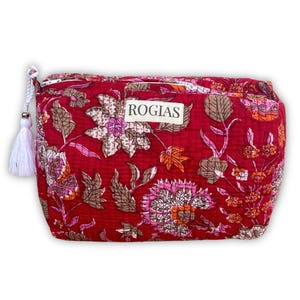 Op de afbeelding: Een rode make-uptas met bloemenprint en een witte kwastritstrekker. De tas heeft een herhalend patroon van roze, oranje en bruine bloemen en bladeren. Het woord "ROGIAS" is gedrukt op een wit label.