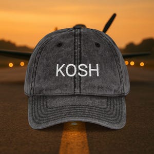 May include: A distressed gray baseball cap with the word "KOSH" embroidered in white block letters. The cap has a curved brim and a classic six-panel design. The background shows an airplane on a runway at sunset.
