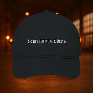 Puede incluir: Gorra de béisbol negra con el texto bordado "I can land a plane" en blanco. La gorra tiene una visera curva y una corona estructurada. El fondo está borroso, con tonos cálidos.