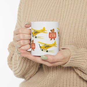 Puede incluir: Taza de cerámica blanca con aviones Piper Cub amarillos y un oso de peluche sosteniendo un letrero que dice "Piper Cub". La taza es de tamaño estándar y tiene un acabado brillante.