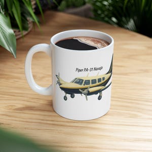 Op de afbeelding: Witte keramische mok met een zwart handvat, gevuld met donkere koffie. De mok heeft een gele en zwarte illustratie van een Piper PA-31 Navajo vliegtuig, met de tekst "Piper PA-31 Navajo".