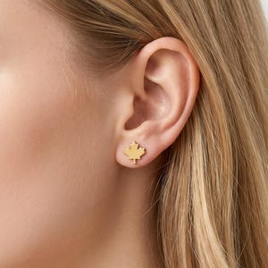 May include: Gold-colored maple leaf stud earrings. The earrings are small, with a detailed leaf design. The earrings are shown on an ear, highlighting their size and shape. The earrings are a simple, elegant accessory.