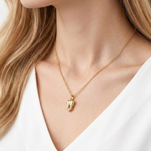 May include: A gold-colored necklace featuring a pendant shaped like a tooth. The tooth pendant has a small, clear stone and is suspended from a delicate gold chain. The necklace is worn against a white top.