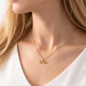 May include: A gold-colored necklace with an infinity symbol pendant. The pendant is a small, stylized figure eight. The necklace is worn against a white top.