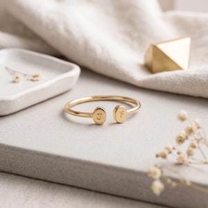 May include: A gold-toned open cuff bracelet featuring two small oval-shaped terminals at the ends. Each terminal is engraved with a single letter, specifically the uppercase letter J on one side and the lowercase letter f on the other side.