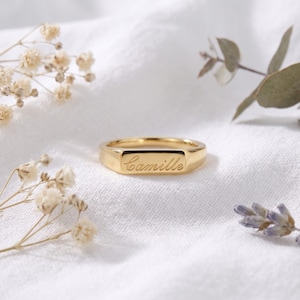 May include: A gold-toned metal ring featuring a flat, rectangular face engraved with the name Camille in a cursive script. The ring has a polished, smooth finish and a simple band design, presented as a singular piece of jewelry.