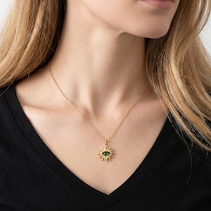 May include: A gold-colored necklace featuring a pendant with an eye design. The eye has a green center and is surrounded by sun-like rays. The necklace is worn against a black top.