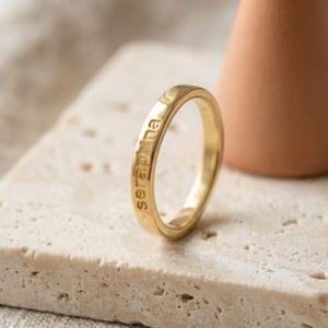 May include: A polished gold-toned band ring featuring the name Seraphina engraved in lowercase letters along the outer surface. The ring has a smooth, rounded profile and a consistent width, showcasing a clean and simple metallic design.