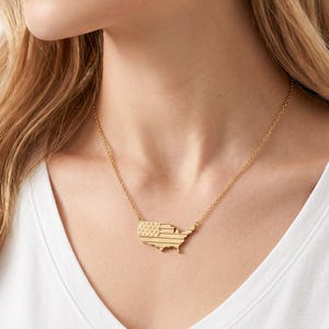 May include: A gold-toned necklace featuring a pendant shaped like the map of the United States. The surface of the map pendant is textured with a pattern resembling the American flag, including a section of small raised dots and horizontal lines.